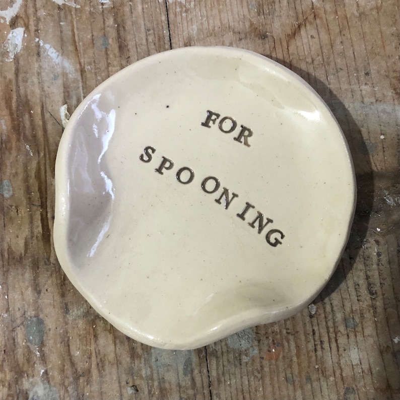 Op de afbeelding: Een wit keramisch schaaltje met een licht onregelmatige vorm. Het schaaltje heeft de woorden "FOR SPOONING" erop geschreven in gouden letters.
