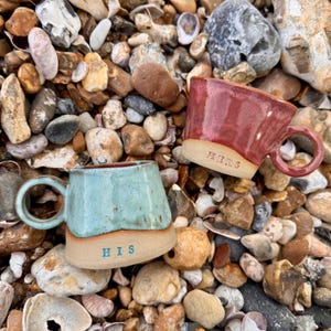 May include: Two handcrafted ceramic mugs, one teal and one red, sit on a bed of pebbles. The teal mug has "HIS" inscribed, while the red mug has "HERS". Both mugs feature a rustic design with rounded handles, perfect for a cosy drink.