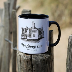 May include: White ceramic mug with a black rim and handle. The mug features a black and white illustration of a building with the text "The Sloop Inn - Circa 1312" printed on the front.