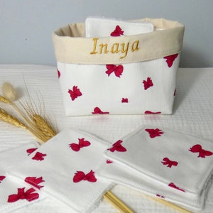 Puede incluir: Una cesta de algodón blanco con lazos rojos impresos en ella. La cesta tiene una parte superior de color crema con el nombre "Inaya" bordado en hilo dorado. La cesta está llena de almohadillas de algodón blanco para desmaquillar con lazos rojos impresos en ellas.