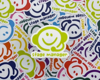 May include: A collection of colorful stickers with a smiling face design and the text "stage manager." The stickers feature a variety of colors, including green, blue, red, and purple, with white text and outlines.