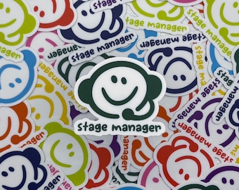 May include: A collection of colorful stickers with a smiling face design and the words "stage manager". The stickers are in various colors, including green, blue, red, and yellow. The stickers are scattered across a white surface.