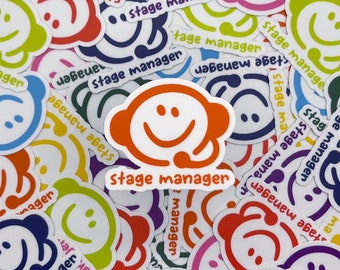 May include: A collection of colorful stickers with a smiling face design and the words "stage manager." The stickers are in various colors, including orange, green, blue, and red. The stickers are scattered across a white surface.