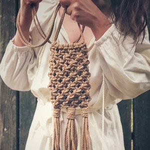 May include: A small, brown, macrame bag with long tassels hanging from the bottom. The bag is being held by a person wearing a white shirt.
