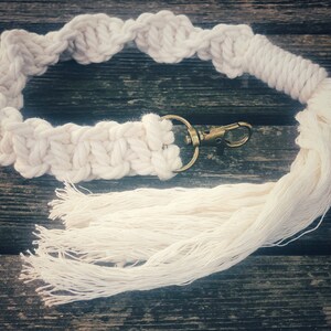 Peut inclure: Un porte-clés en macramé blanc avec un anneau de clé en laiton et un long pompon. Le porte-clés est posé sur une surface en bois.