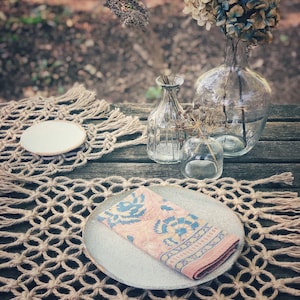 May include: A table setting with a woven placemat, a white plate, a pink and blue patterned napkin, and two glass vases with dried flowers.