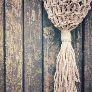 May include: A close-up of a natural fiber rope macrame wall hanging with a long tassel. The macrame is hanging against a weathered wooden background.