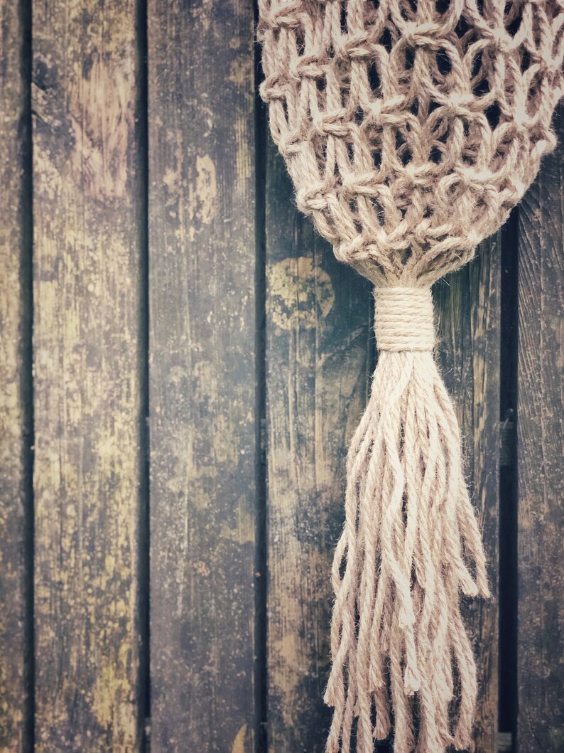 May include: A close-up of a natural fiber rope macrame wall hanging with a long tassel. The macrame is hanging against a weathered wooden background.
