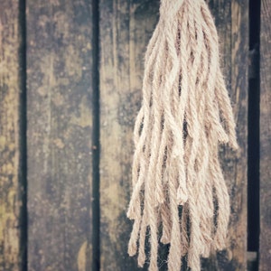 May include: A close-up of a natural fiber rope tassel hanging against a weathered wooden background. The tassel is made of thick, natural-colored rope and has a frayed, uneven texture.