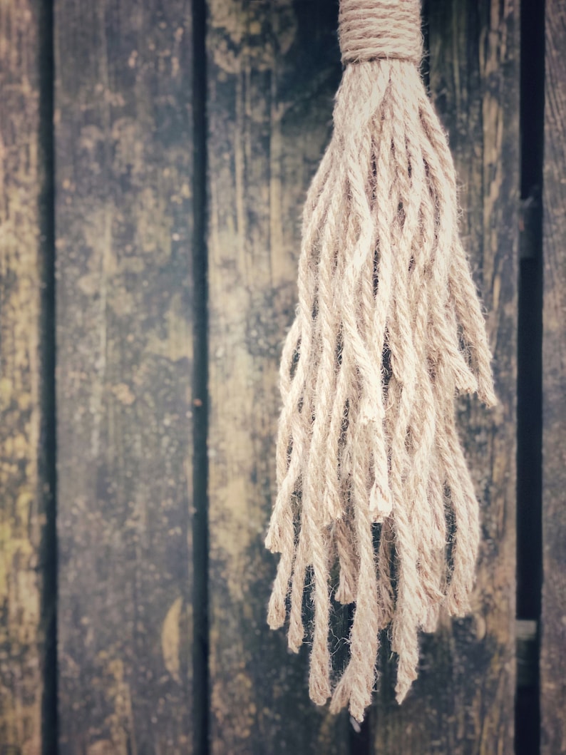 May include: A close-up of a natural fiber rope tassel hanging against a weathered wooden background. The tassel is made of thick, natural-colored rope and has a frayed, uneven texture.