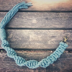 May include: A teal blue rope keychain with a gold metal clasp. The keychain is made of braided rope and is lying on a wooden surface.