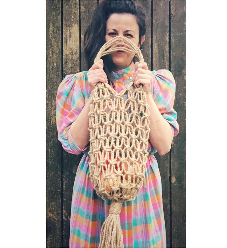 May include: A woman in a pink and blue plaid dress holds a large, natural fiber, macrame bag in front of her face. The bag has a long tassel.