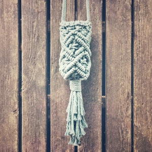 Puede incluir: Un colgador de plantas de macramé hecho a mano en un suave color verde apagado. El intrincado diseño tejido presenta una forma bulbosa central con una borla en la parte inferior. El colgador se suspende con un cordón a juego.
