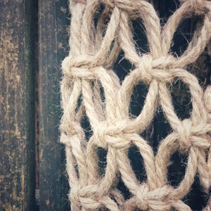 May include: Close-up of a beige macrame wall hanging with a knotted rope design. The macrame is hanging against a dark wood background.