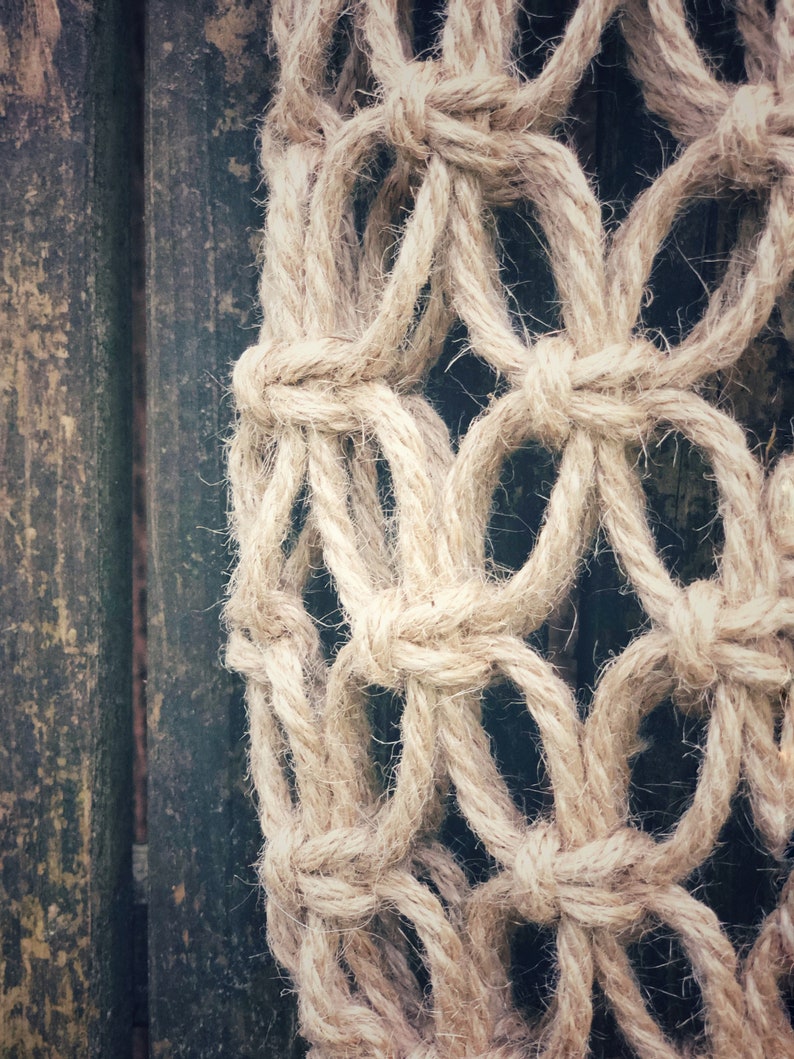 May include: Close-up of a beige macrame wall hanging with a knotted rope design. The macrame is hanging against a dark wood background.