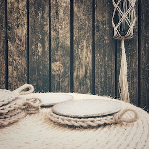 May include: A rustic scene featuring a bottle in a macrame holder, hanging against a weathered wooden backdrop. Two round coasters and a knitted item rest on a textured surface, all in neutral tones.