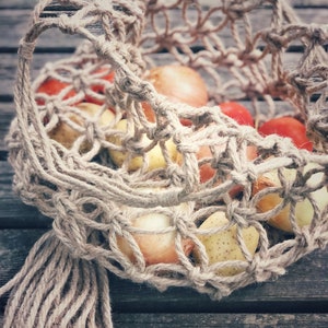 May include: A close-up of a brown, woven string bag filled with red and yellow produce. The bag has a long, knotted fringe hanging from the bottom.