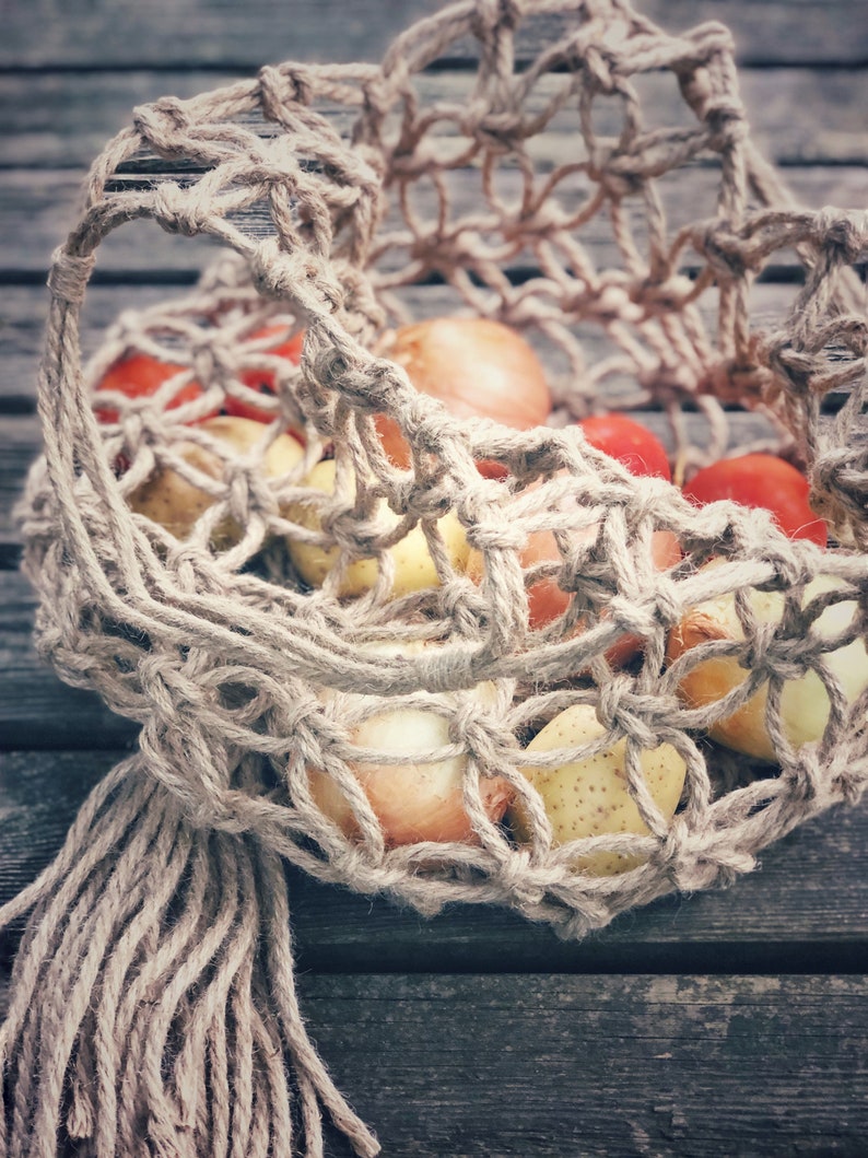 May include: A close-up of a brown, woven string bag filled with red and yellow produce. The bag has a long, knotted fringe hanging from the bottom.