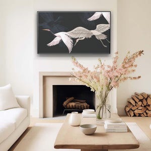 May include: A framed artwork featuring white cranes in flight against a dark background hangs above a fireplace. The artwork is the focal point of a living room with a white sofa, wooden coffee table, and decorative flowers.