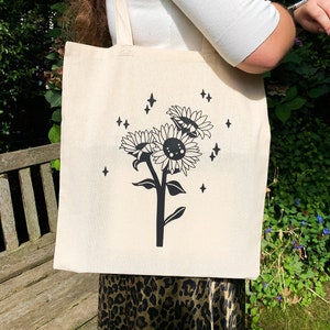 K&ouml;nnte beinhalten: Eine wei&szlig;e Canvas-Tragetasche mit schwarzen Henkeln und einer schwarz-wei&szlig;en Illustration einer Sonnenblume mit einem l&auml;chelnden Gesicht und Sternen. Die Sonnenblume ist von einem Ring aus Sternen umgeben.