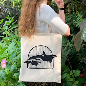 May include: A white canvas tote bag with a black graphic of two orcas swimming under an archway. The orcas are silhouetted against a white background.