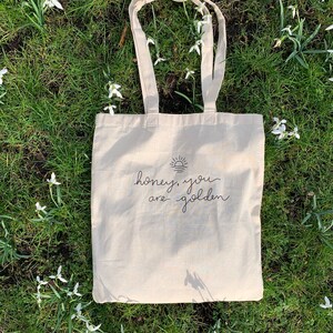 May include: A beige tote bag with black text that reads "honey, you are golden" and a simple sun graphic. The bag is lying on a bed of green grass with white flowers.