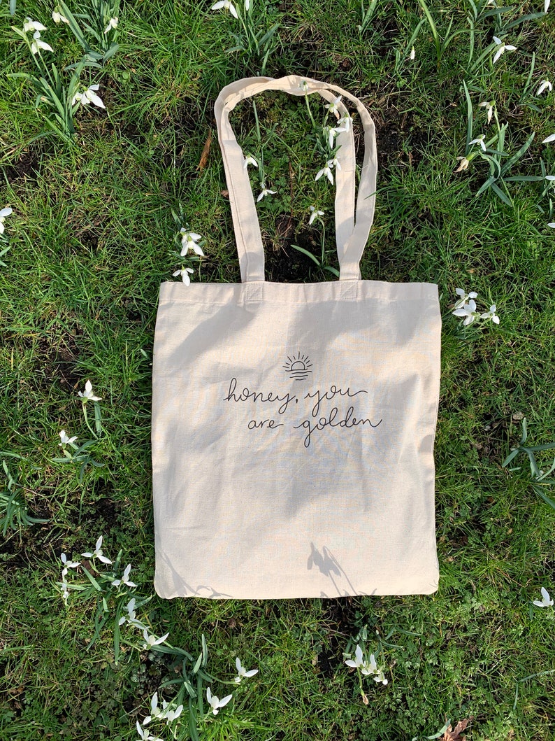 May include: A beige tote bag with black text that reads "honey, you are golden" and a simple sun graphic. The bag is lying on a bed of green grass with white flowers.