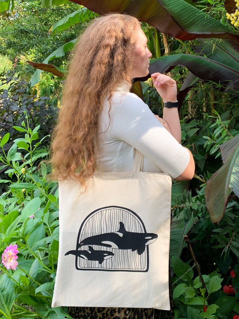 Puede incluir: Una bolsa de lona blanca con un gr&aacute;fico negro de dos orcas nadando bajo un arco. Las orcas est&aacute;n silueteados sobre un fondo blanco.