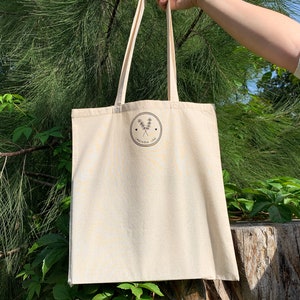 Puede incluir: Una bolsa de tela beige con un logotipo negro que dice "Lavender Love" con un peque&ntilde;o dise&ntilde;o floral y dos corazones. La bolsa est&aacute; siendo sostenida por una mano frente a un fondo verde frondoso.