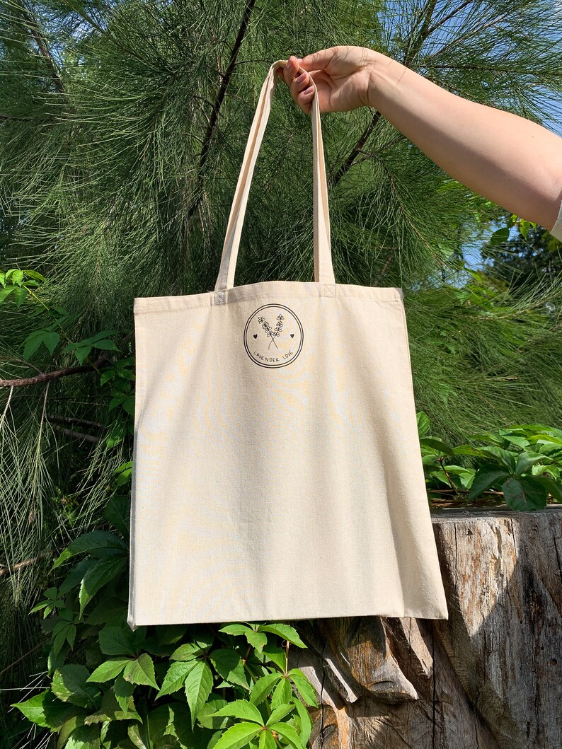 Puede incluir: Una bolsa de tela beige con un logotipo negro que dice "Lavender Love" con un peque&ntilde;o dise&ntilde;o floral y dos corazones. La bolsa est&aacute; siendo sostenida por una mano frente a un fondo verde frondoso.