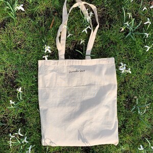 May include: A white canvas tote bag with the text "Lavender Love" printed on the front. The bag is lying on a bed of green grass with white flowers.