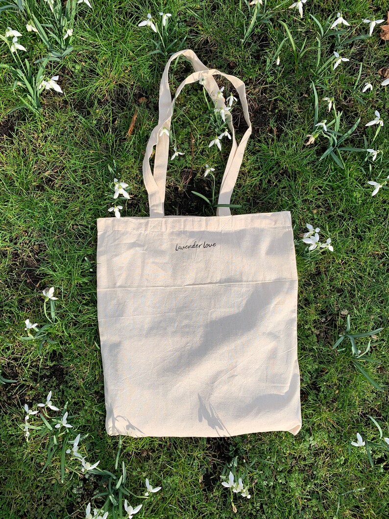 May include: A white canvas tote bag with the text "Lavender Love" printed on the front. The bag is lying on a bed of green grass with white flowers.
