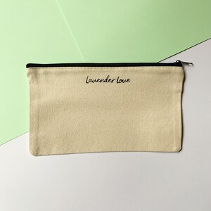 Op de afbeelding: Een beige canvas etui met een zwarte rits en zwarte bies. Het etui heeft de tekst "lavender love" in het zwart op de voorkant gedrukt.