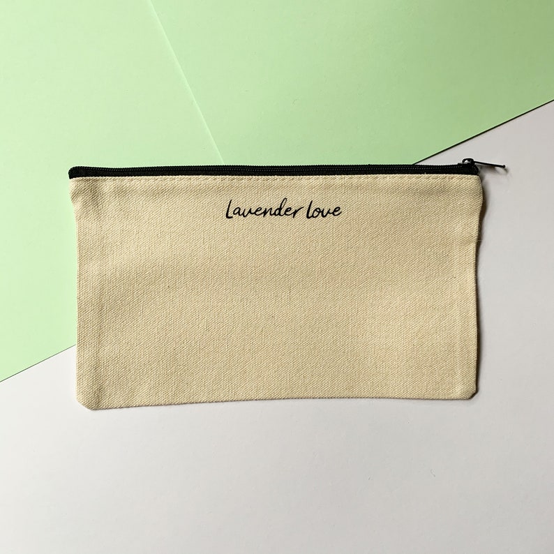 Op de afbeelding: Een beige canvas etui met een zwarte rits en zwarte bies. Het etui heeft de tekst "lavender love" in het zwart op de voorkant gedrukt.