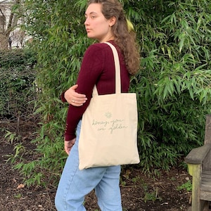 May include: A person is holding a beige tote bag with a green sun and the text "honey, you are golden" printed on it.