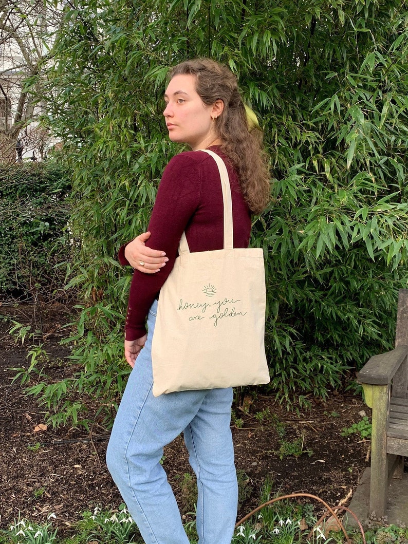 May include: A person is holding a beige tote bag with a green sun and the text "honey, you are golden" printed on it.