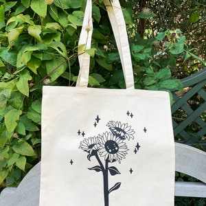 K&ouml;nnte beinhalten: Eine wei&szlig;e Canvas-Tragetasche mit einem schwarzen Sonnenblumen-Design. Die Sonnenblumen haben l&auml;chelnde Gesichter und sind von kleinen Sternen umgeben.