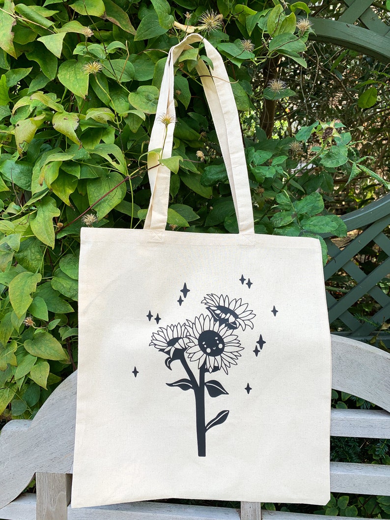 K&ouml;nnte beinhalten: Eine wei&szlig;e Canvas-Tragetasche mit einem schwarzen Sonnenblumen-Design. Die Sonnenblumen haben l&auml;chelnde Gesichter und sind von kleinen Sternen umgeben.
