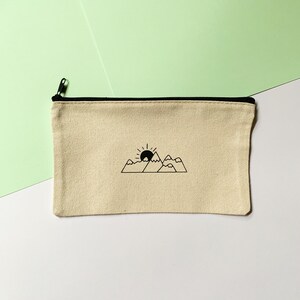 Op de afbeelding: Een beige canvas toilettas met een zwarte rits en een zwarte lijntekening van een zon die ondergaat achter bergen.