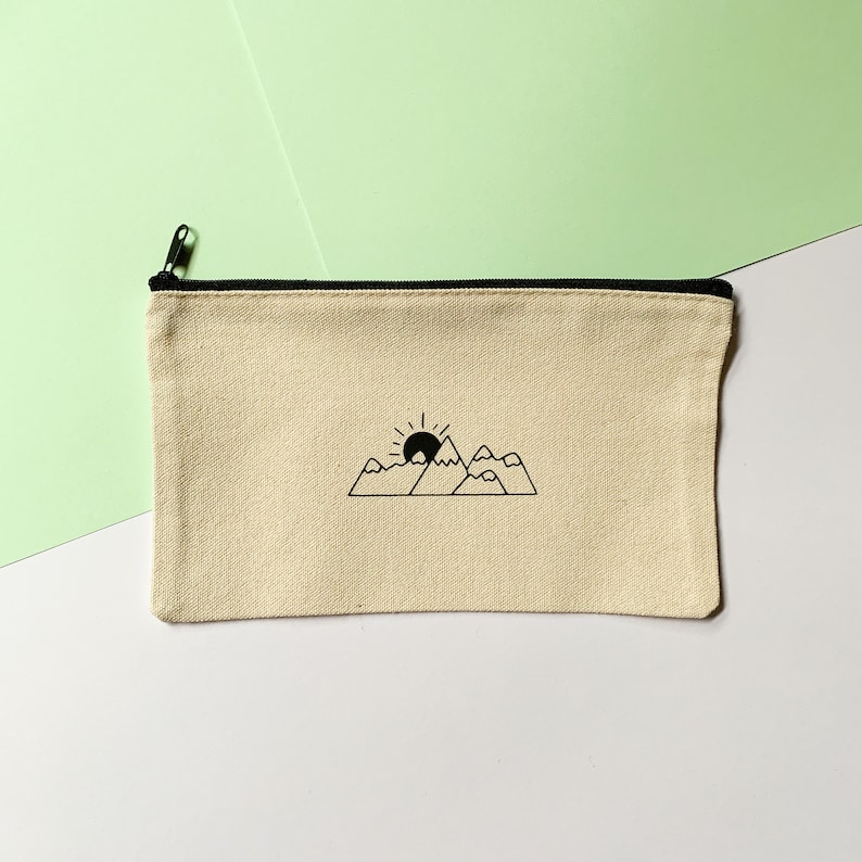 Op de afbeelding: Een beige canvas toilettas met een zwarte rits en een zwarte lijntekening van een zon die ondergaat achter bergen.