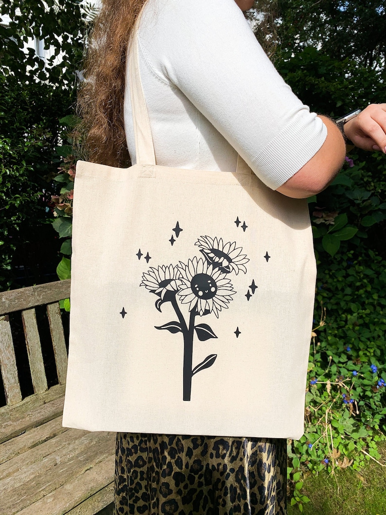 K&ouml;nnte beinhalten: Eine wei&szlig;e Canvas-Tragetasche mit schwarzen Henkeln und einer schwarz-wei&szlig;en Illustration einer Sonnenblume mit einem l&auml;chelnden Gesicht und Sternen. Die Sonnenblume ist von einem Ring aus Sternen umgeben.