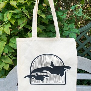 Puede incluir: Una bolsa de lona blanca con asas negras y un gr&aacute;fico negro de dos orcas nadando en un dise&ntilde;o de ola. Las orcas est&aacute;n silueteados sobre un fondo blanco.
