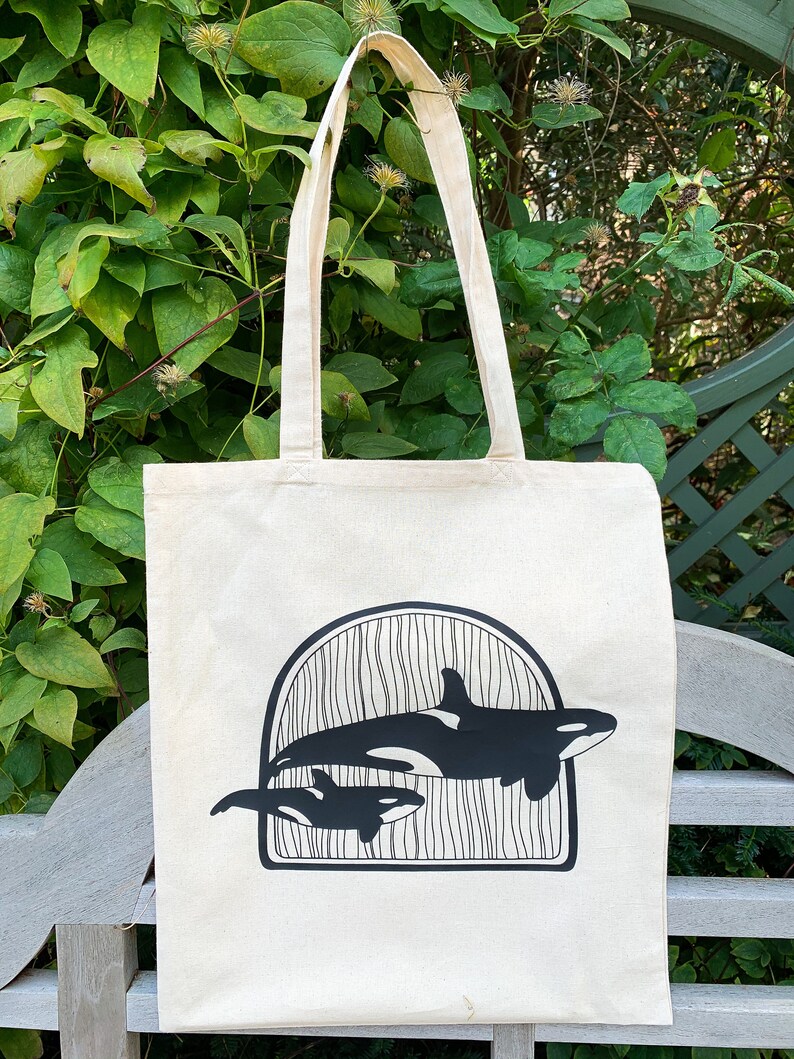 Puede incluir: Una bolsa de lona blanca con asas negras y un gr&aacute;fico negro de dos orcas nadando en un dise&ntilde;o de ola. Las orcas est&aacute;n silueteados sobre un fondo blanco.