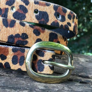 May include: A brown and black leopard print belt with a gold metal buckle. The belt is folded in half and the buckle is on the right side of the image.