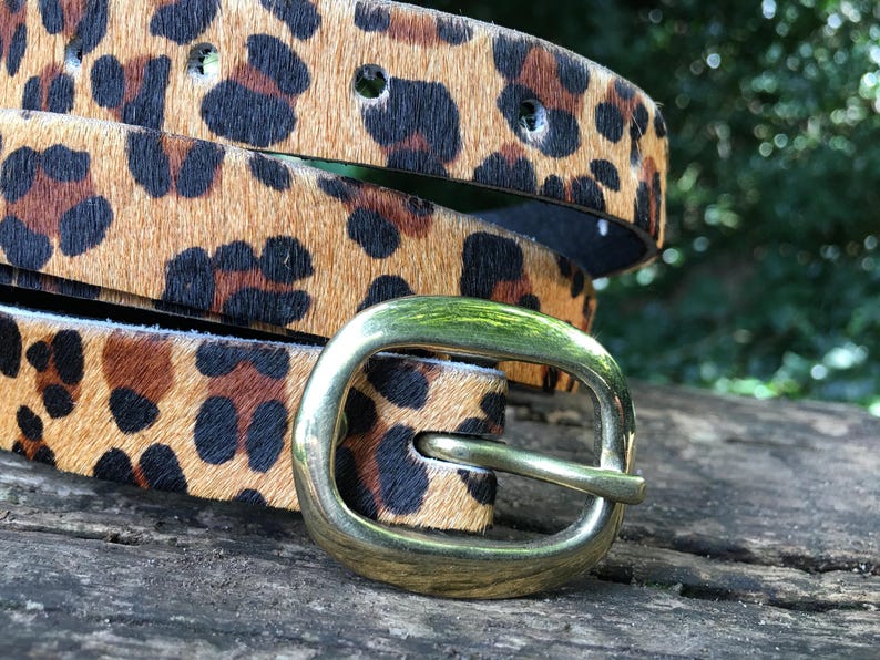 May include: A brown and black leopard print belt with a gold metal buckle. The belt is folded in half and the buckle is on the right side of the image.