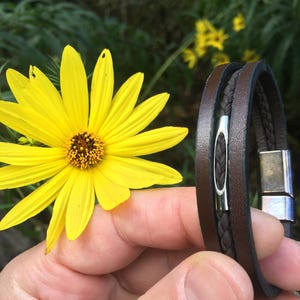 May include: A brown leather bracelet with a braided center section and a silver magnetic clasp. The bracelet is held near a bright yellow flower with a brown center, set against a blurred green background.