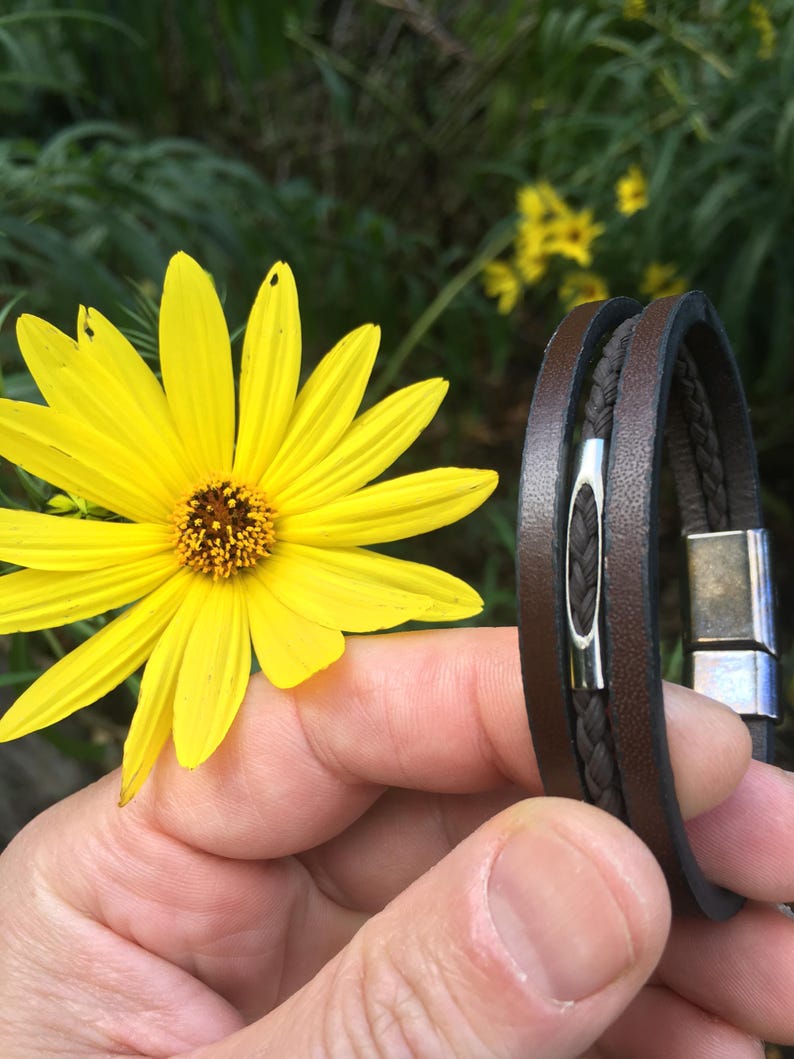 May include: A brown leather bracelet with a braided center section and a silver magnetic clasp. The bracelet is held near a bright yellow flower with a brown center, set against a blurred green background.