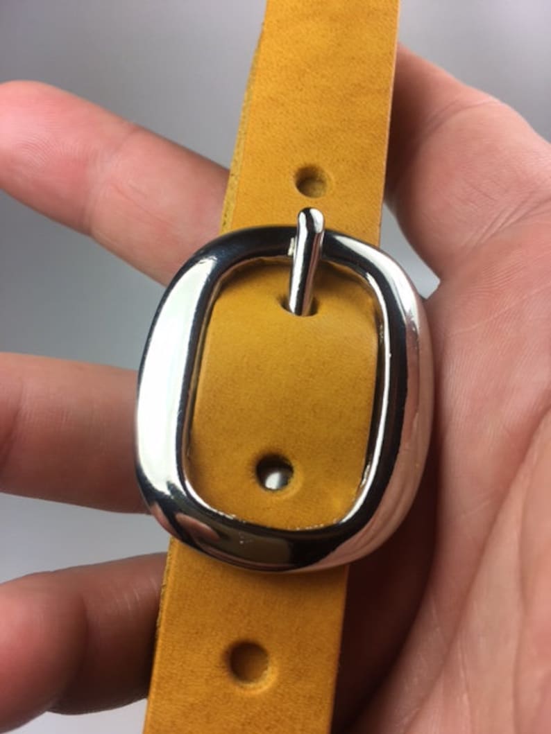 Puede incluir: Primer plano de una correa de cuero amarilla con una hebilla plateada. La hebilla tiene un acabado liso y pulido y un dise&ntilde;o sencillo.