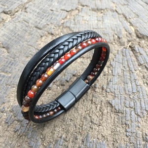 May include: Black leather bracelet featuring braided and smooth bands, with small, round, orange and brown beads. The bracelet has a magnetic clasp. A stylish accessory.