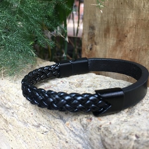 Puede incluir: Un brazalete de cuero negro con un diseño trenzado y un cierre de metal.